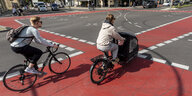 Ein Mensch fährt auf einem sportlichen Rad, eine andere Person auf einem E-Lastenrad auf einem rot markierten Fahrradweg über eine Kreuzung
