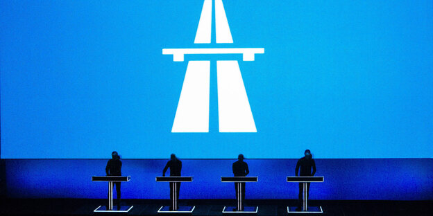 Im Hintergrund das blaue Autobahnzeichen, im Vordergrund die vier Bandmitglieder von Kraftwerk