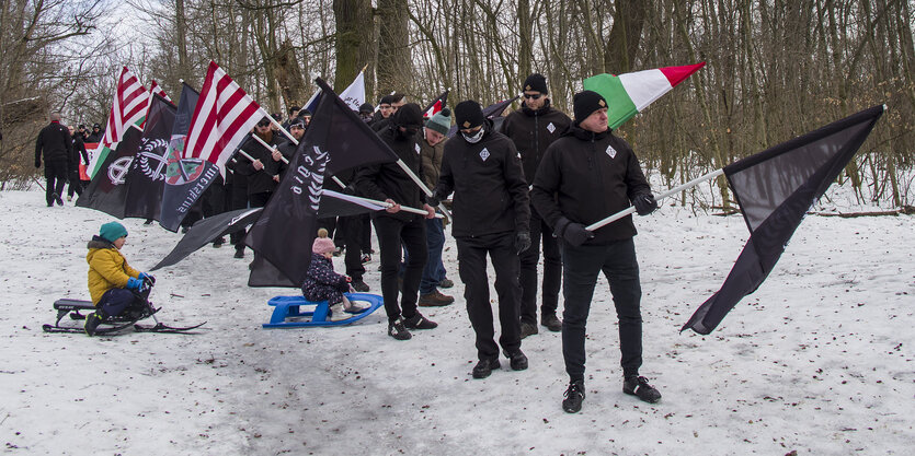 Zwei kleine Kinder fahren auf Schlitten zwischen einer Parade von uniformierten Fahnenträgern hindurch