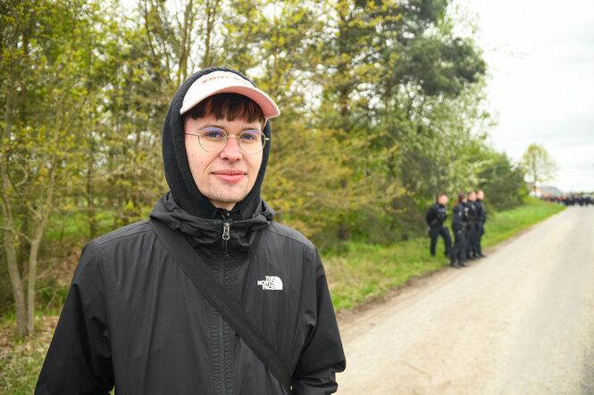 Der linke Aktivist steht auf einem Feldweg, im HIntergrund die Polizei