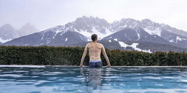 Ein Mann stützt sich auf den beckenrand eines Schwimbeckens und blickt auf ein Alpenpanorama