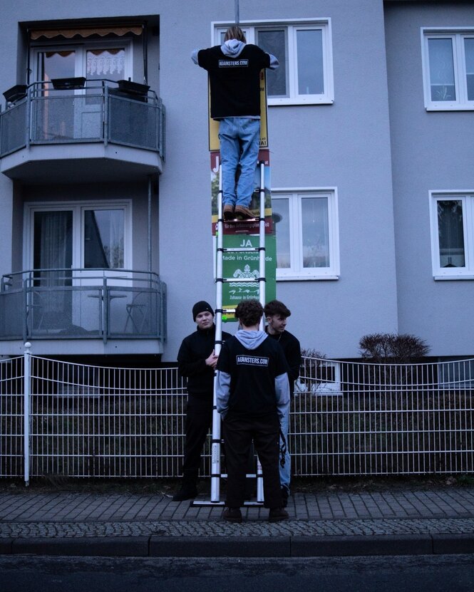 Beim Plakatehängen halten drei Menschen eine Leiter, ein Mensch steht darauf