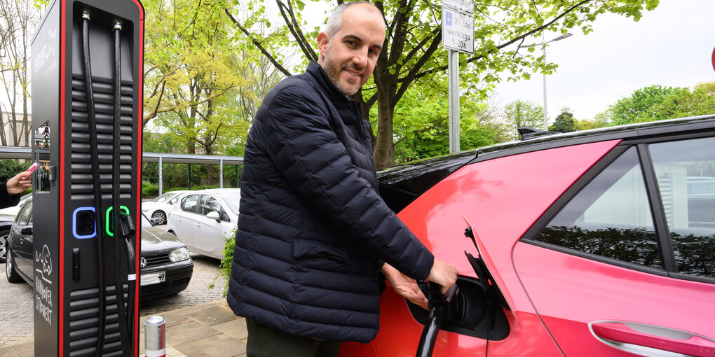 Hannovers Oberbürgermeister Belit Onay steht an einer Ladesäule für Elektroautos und "betankt" ein rotes Auto.