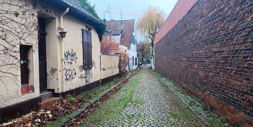 Gepflasterte Gasse, rechts eine hohe Klinkermauer, rechts flache Häuser