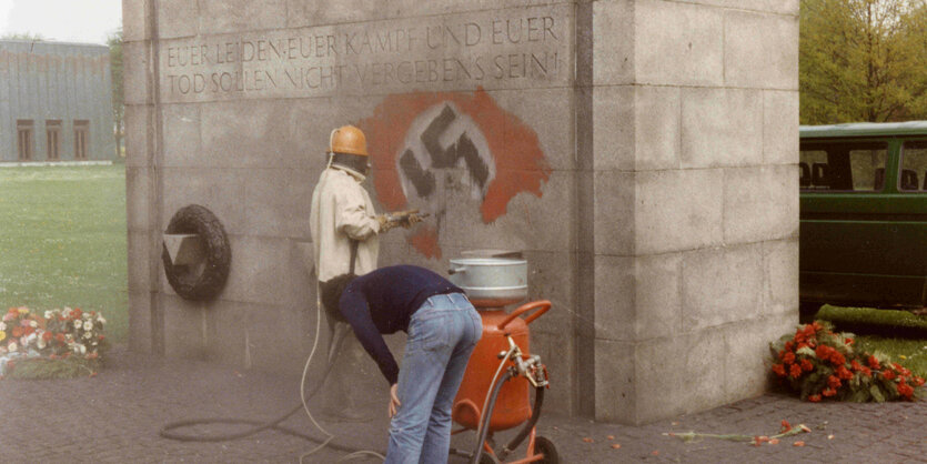 Zwei Menschen reinigen ein Mahnmal mit einem Dampfstrahler von einem Hakenkreuz