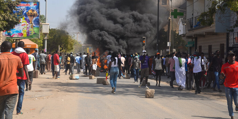 Viele Menschen auf einer Straße in Dakar. Im Hintersgrund steigen schwarze Rauchschwaden auf