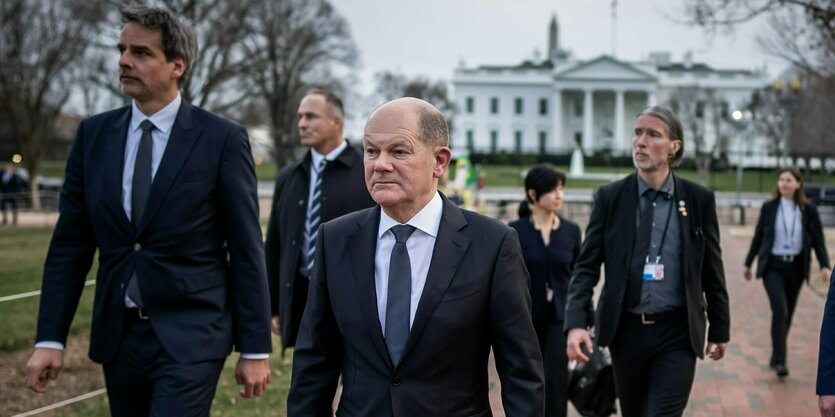 Bundeskanzler Olaf Scholz (SPD) läuft mit einer Gruppe von Mitarbeitern und ernstem Gesicht dem Betrachtert entgegen, im Hintergrund ist das Weiße Haus zu sehen.