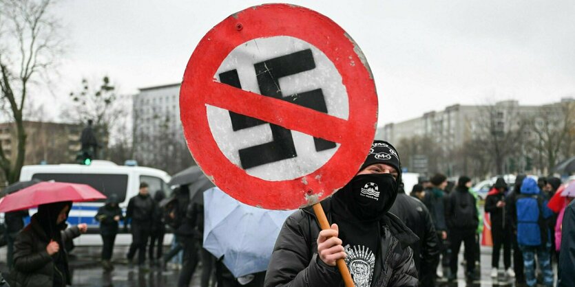 Vermummter Demonstrant hält ein rundes Schild in Form eines Stoppschilds mit durchgestrichenem Hakenkreuz hoch. Im Hintergrund Menschen mit Regenschirmen und Regenwetter.