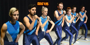 DarstellerInnen in Gymnastikkleidung singen in Mikrofone