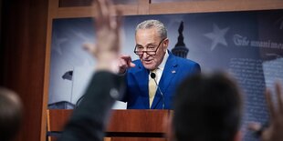 Chuck Shumer steht an einem Rednerpult und zeigt mit dem Fingerauf einen Menschen, der seinen Arm gehoben hat