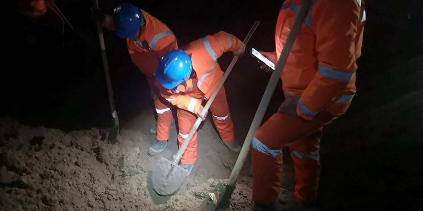 Menschen mit orangefarbenen Arbeitsanzügen und blauen Helmen schaufeln Erde