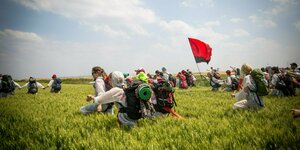 Eine Aktivistengruppe mit weißen Schutzanzügen läuft mit schwarz-roter Fahne durch ein grünes Getreidefeld