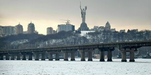 Kyjiw im Winter. Es liegt Schnee auf einer freien Fläche. Es ist nicht erischtlich, ob es ein zugefrorener See oder eine Wiese ist. Auf der Fläche Menschen, die etwas sammeln. Dahinter eine lange brücke. Hinter der Brücke Häuser und ein riesiger weiblicher Engel, eine Statue oder ein Denkmal. Der Engel hebt die Arme und hat ein Schwert dabei.