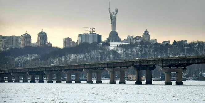 Kyjiw im Winter. Es liegt Schnee auf einer freien Fläche. Es ist nicht erischtlich, ob es ein zugefrorener See oder eine Wiese ist. Auf der Fläche Menschen, die etwas sammeln. Dahinter eine lange brücke. Hinter der Brücke Häuser und ein riesiger weiblicher Engel, eine Statue oder ein Denkmal. Der Engel hebt die Arme und hat ein Schwert dabei.