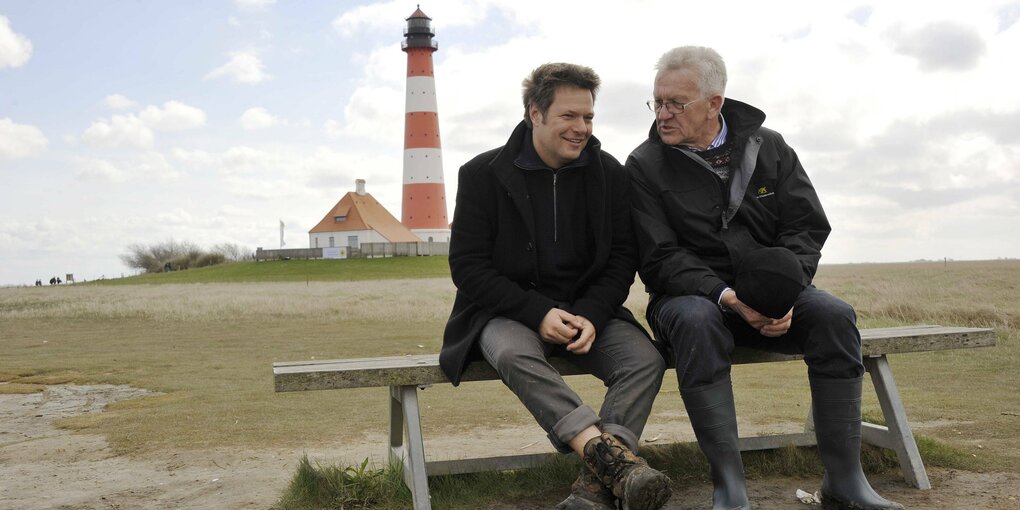 Winfried Kretschmann und Robert Habeck auf einer Bank vor einem Leuchtturm.