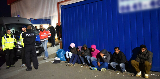 Menschen sitzen vor einem Container, links im Bild Polizei.