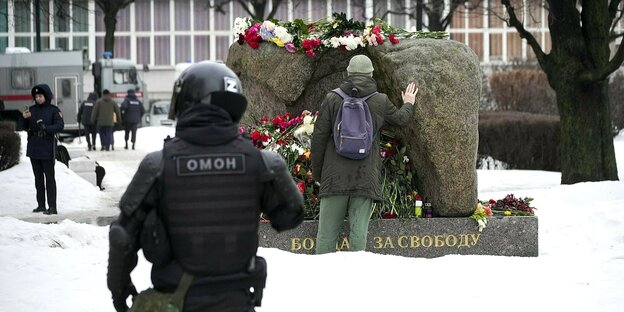 Ein Mann steht an einem Denkmal, auf dem viele Blumen liegen. Im Vordergrund ist ein Polizeibeamte von hinten zu sehen