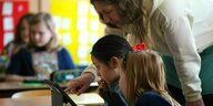 Eine Lehrerin beugt sich über ihre Schülerinnen, um ihnen etwas am Tablet zu zeigen