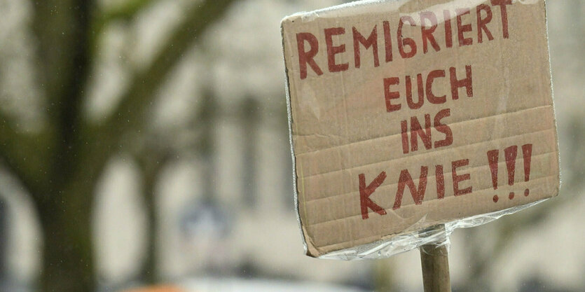 Menschen stehen im Regen, einer hält ein Pappschild hoch. Dort steht: Remigriert euch ins Knie