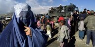 Eine Frau, die eine Burka trägt steht einer Menschenmenge und patrouiliienden afghanischen Polizisten