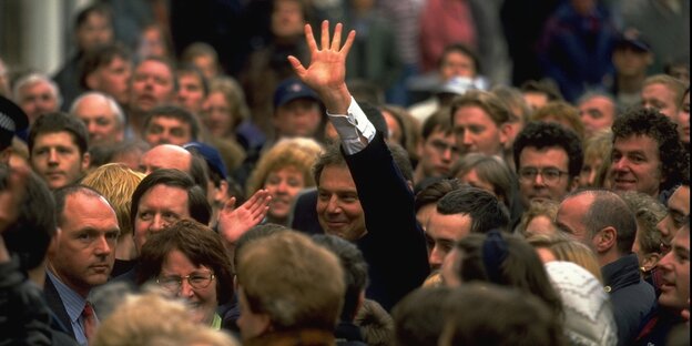 Tony Blair in einer Menschenmenge. Man sieht nur seinen Kopf und seine linke Hand, die er in die Luft streckt. Blair lächelt.