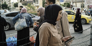 Eine Frau mit Kopftuch tippt auf ihrem Handy, im Hintergrund weitere Frauen und ein paar Männer mit Einkaufstaschen auf einem zentralen belebten Platz in Ramallah