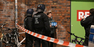 eine Mensch steht vor einer Hauswand und wird von Polizist:innen fotografiert