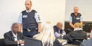 Angeklagte sitzen im Gericht. Neben ihnen stehen Polizisten.