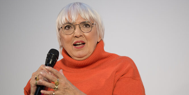 Claudia Roth im orangen Pullover und Mikrofon in der Hand