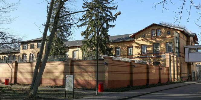 Ein Backsteingebäude mit vergitterten Fenstern, das von einer hohen Steinmauer und Stacheldrahtzaun umgeben ist. Davor stehen Bäume.