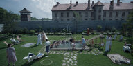 Ein Wachturm und ein Konzentrationslager, umschlossen von einer Mauer, davor baden Kinder in einem Schwimmbad, gekleidete Erwachsene auf einer grünen Wiesen