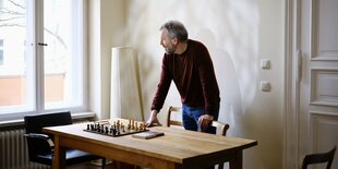 Ein älterer Mann im roten Pullover steht leicht gebeugt an einem Tisch mit Schachbrett und schaut aus dem Fenster, das Licht wirft einen Shhatten auf die Wand