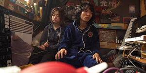zwei junge Chinesen in Trainingsanzügen sitzen in einem sehr engen, vollgestellten Zimmer