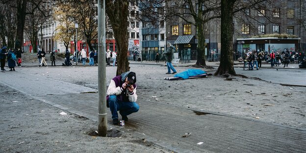 Ein Mann kniet mit Fotoapparat in der Hand, ein Mensch liegt im Schlafsack auf grauer Erde