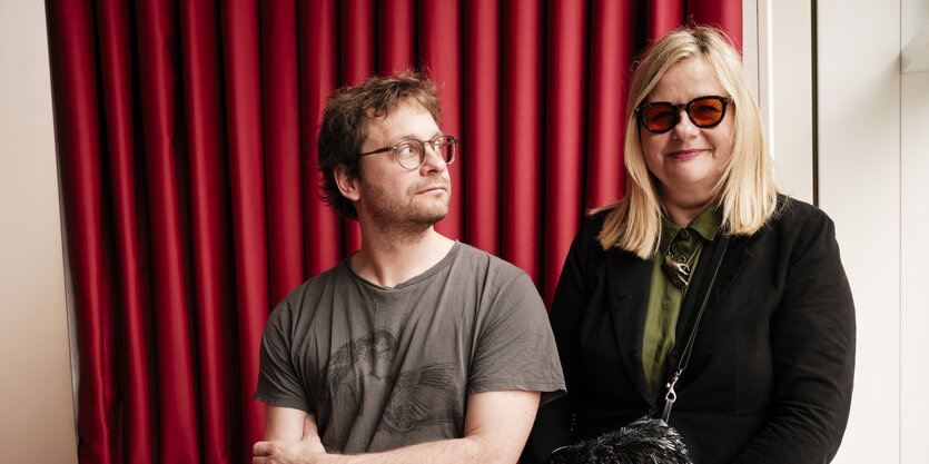 Ein Mann und eine Frau, er trägt eine Brille und schaut nach rechts zu ihr, sie ist blond und lächelt