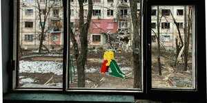 Blick aus dem Fenster auf einen zerstörten Wohnblock nach einem Bombenangriff. Vor dem Fenster steht eine bunte Kinderrutsche