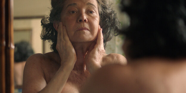 Eine ältere Frau steht nackt vor dem Spiegel und betrachtet sich