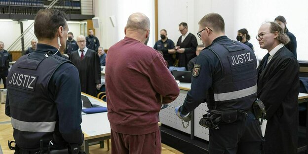 Gerichtssaal mit einem Angeklagten in weinrotem Outfit, bneben ihm stehen zwei Polizisten, einer von ihnen ist mit Handschellen an den Angeklagten gekettet