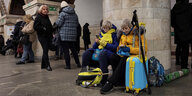 Zwei ältere Frauen mit blau-gelben Koffern und in blau-gelber Kleidung sitzen in einem U-Bahnhof in Kyiv und schauen auf ihre Handys
