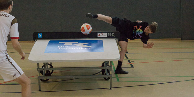 zwei Teqball-Spieler*innen an einer Tischtennisplatte; eine der Personen schießt gerade einen Ball über die Platte - ein Bein auf dem Boden, eines fast waagerecht zum Tisch, der Oberkörper zu Seite abgeknickt