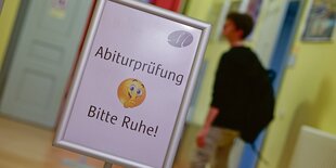 Ein Schüler läuft an einem Schild vorbei, auf dem "Abiturprüfung - Bitte Ruhe" steht