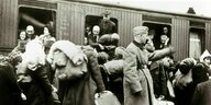 Juden stehen mit Gepäck auf dem Bahnsteig, auf dem ein Waggon steht. Erwachsene und Kinder schauen aus dem Abteilfenster. Ein Mann in Uniform hebt den Arm