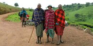 Drei Massais stehen eingewickelt in karierte Decken auf einer unbefestigten Straße, hinter ihnen kommt eine lächelnde Massaifrau. Alle haben Holzstöcke in der Hand
