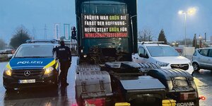 Eine schwarze LKW-Zugmaschine mit Transparent am Führerhaus, daneben steht ein Polizeiauto.