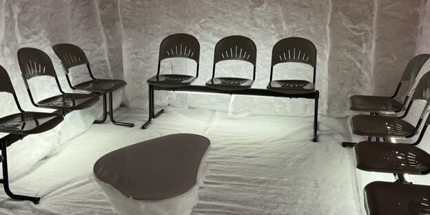 In einem komplett mit weißem Fließ ausgekleideten Raum stehen Metallstühle in Dreierpaaren an den Wänden. In der Raummitte steht ein organisch geformter weißer Tisch