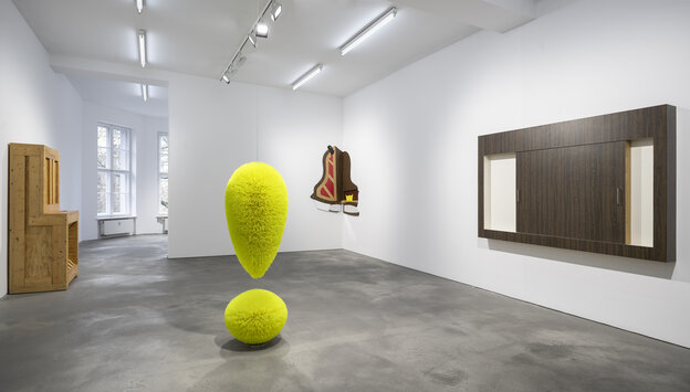 Ausstellungsansicht. In der Raummitte steht ein Ausrufezeichen aus gelben Borsten. Links an der Wand ist eine Transportbox zu sehen, die die Form eines kleinen Klaviers andeutet. In der rechten Raumecke hängt eine flache, aufgefächerte Holzskulotur, die d