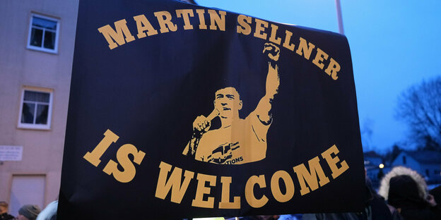 Eine große schwarze Flagge mit gelber Schrift auf der steht: Martin Sellner is welcome und einer Abbildung von ihm - erhobene Faust, er spricht in ein Mikrofon