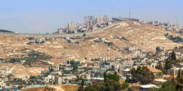Blick auf die Landschaft bei Jerusalem, im Vordergrund ein palästinensischer Ort, Sonne scheint auf sandige Landschaft, eine Mauer trennt Siedlungsbereiche