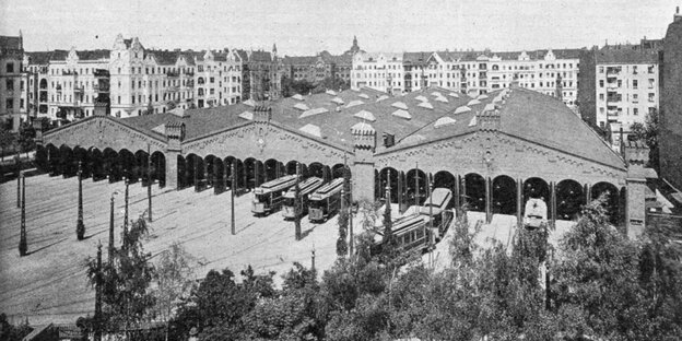 Das schwarz-weiß-Foto zeigt eine große dreigliedrige Halle, davor Schienen und alte Straßenbahnen. Dahinter Wohnhäuser.