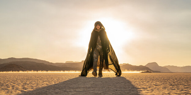 Über den Sand marschiert ein Mann mit Umhang auf den Betrachter zu, hinter ihm steht die Sonne tief